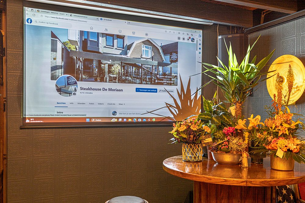 Het beamerscherm in de achterzaal met bloemen ervoor